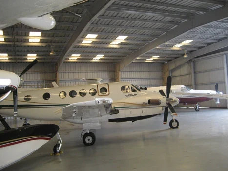 Aircraft parked inside a hangar with exposed steel roof framing and overhead lights