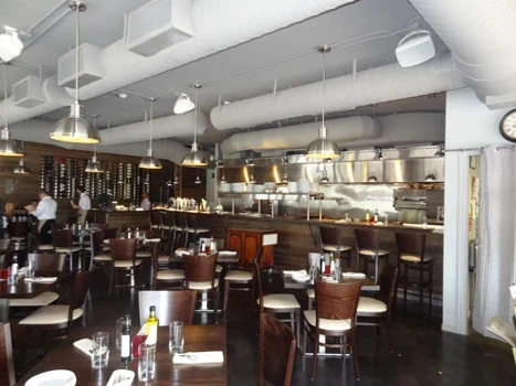 Restaurant dining room with tables, chairs, exposed white ducts, and a bar